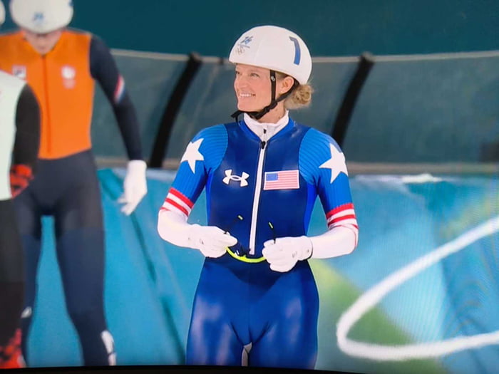 A young female speed skater wearing a blue USA suit and white helmet, standing on an ice rink.
