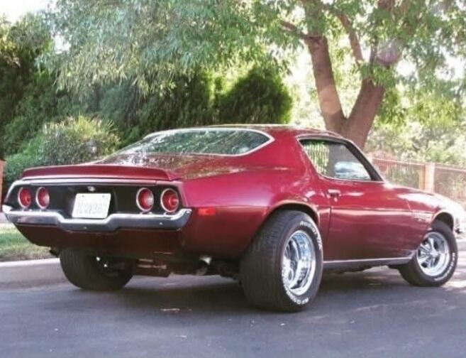 A classic dark red Chevrolet Camaro, second generation, parked outdoors. The car features chrome bumpers, round taillights, and wide chrome wheels with BFGoodrich tires. The background shows green trees and bushes.