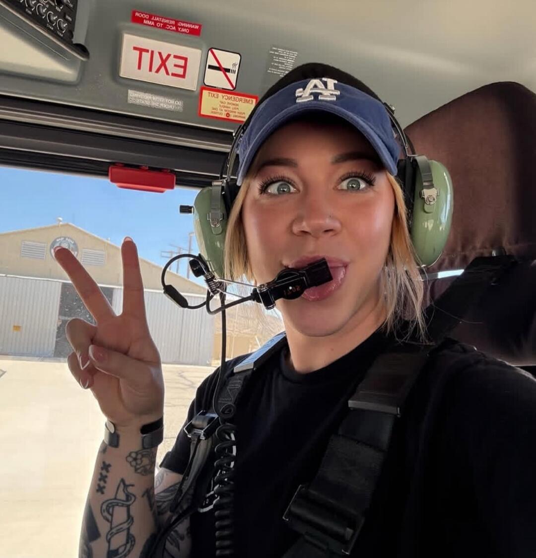 A woman in a helicopter makes a funny face, sticking out her tongue and showing a peace sign. Signs visible inside the helicopter include 'EXIT' (reversed) and a 'NO SMOKING' symbol.