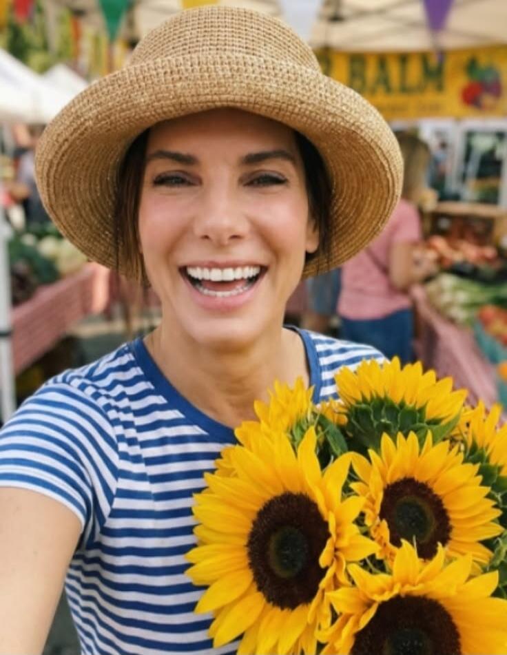 A smiling woman in a straw hat and striped shirt holds a bouquet of sunflowers at an outdoor market. The word 'BALM' is visible on a banner in the background.