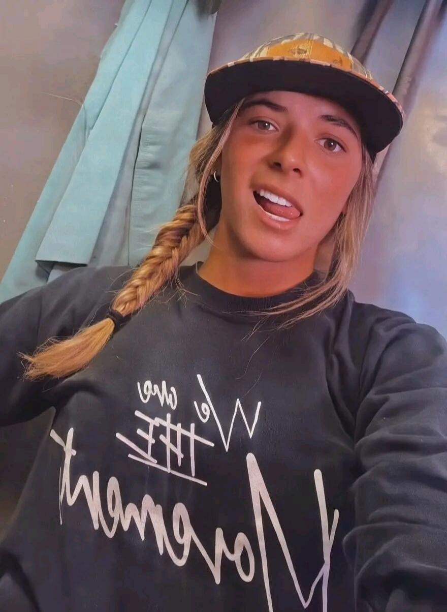 A young woman wearing a cap and a dark sweatshirt, sticking her tongue out playfully. The text on her sweatshirt appears to be 