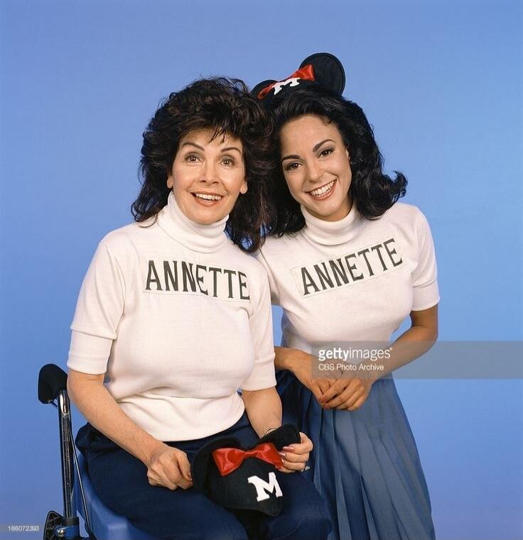 Two women wearing white turtleneck shirts with 'ANNETTE' printed on them. One woman is wearing Mickey Mouse ears, and the other is holding a black hat with an 'M' and a red bow.
