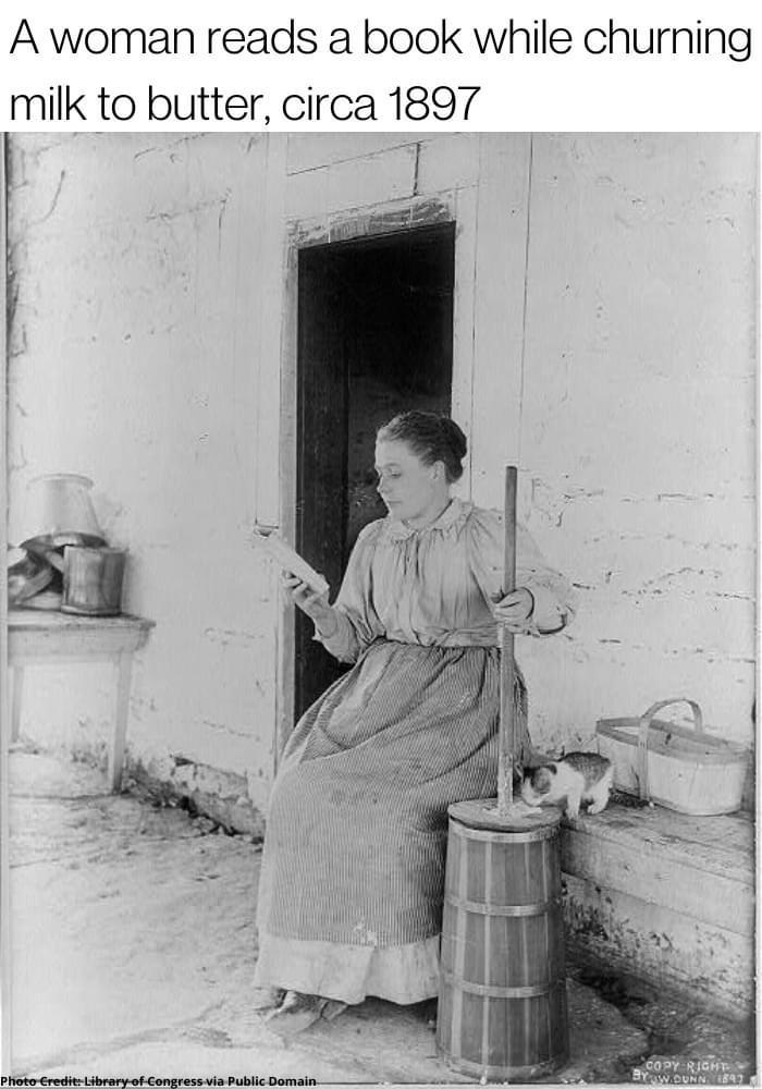 A woman reads a book while churning milk to butter circa 1897