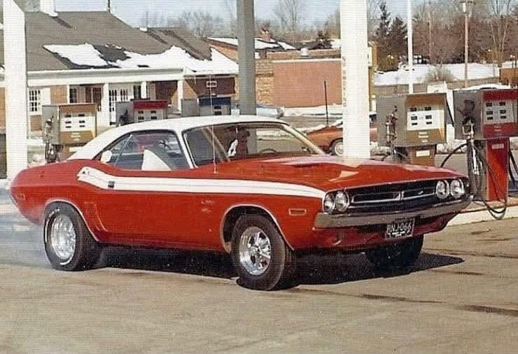 A classic red Dodge Challenger with a white roof and white stripes is performing a burnout, creating smoke from its tires, at a gas station. The license plate reads RNJ-066. Buildings with snow on their roofs are visible in the background.
