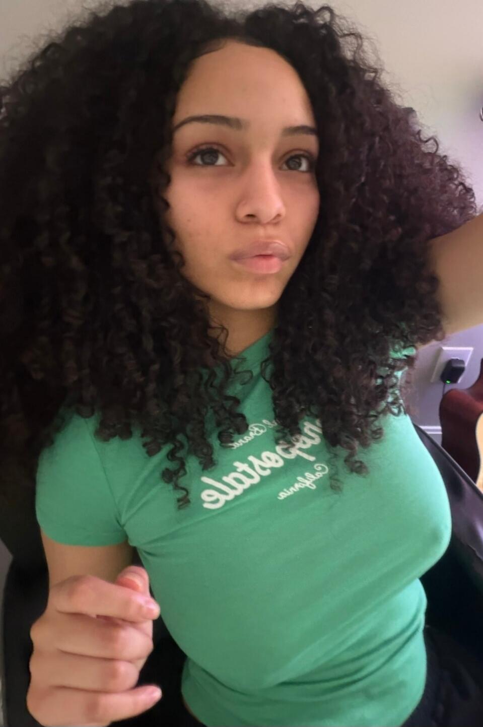 A young woman with dark curly hair is taking a selfie. She is wearing a tight green t-shirt with mirrored text that appears to say 'Aeropostale California'. Her nipples are visible through the fabric of the shirt.