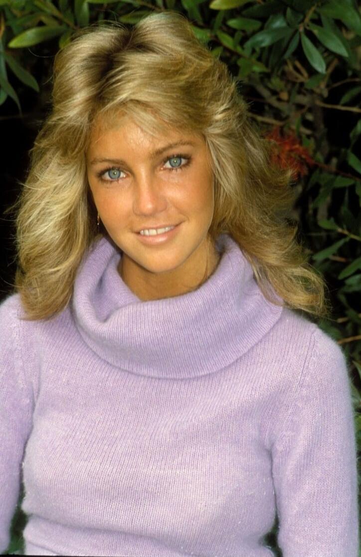 A portrait photo of a smiling woman with blonde hair wearing a lavender turtleneck sweater, outdoors with greenery in the background.