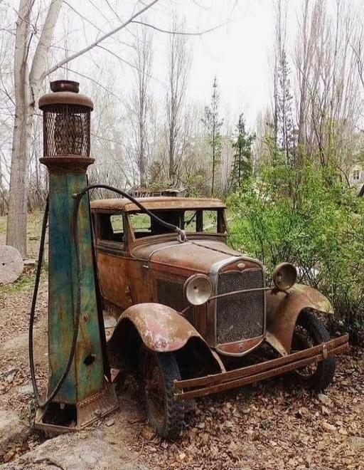 An old rusty vintage car beside a tall, weathered lamp in a wooded, abandoned setting.