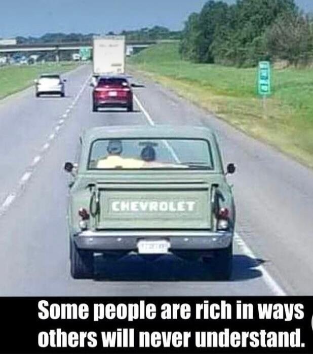 An old green Chevrolet pickup truck is driving on a highway with two people inside. Other vehicles are also on the road. Text on the image says: 