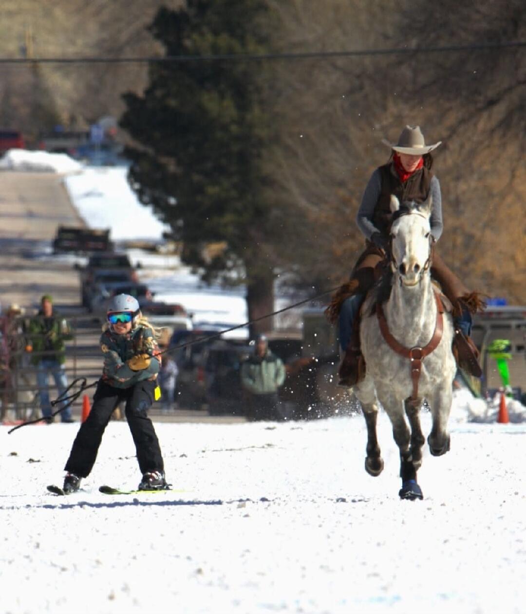 A person on horseback is pulling a child on skis across a snowy landscape. The child is wearing a helmet and goggles, holding onto a rope, while the horse and rider are in motion, kicking up snow.