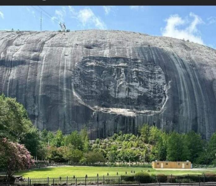 A large granite rock cliff with a large circular carving or weathered pattern resembling a face, set in a park with trees, a lawn, and a small yellow building.