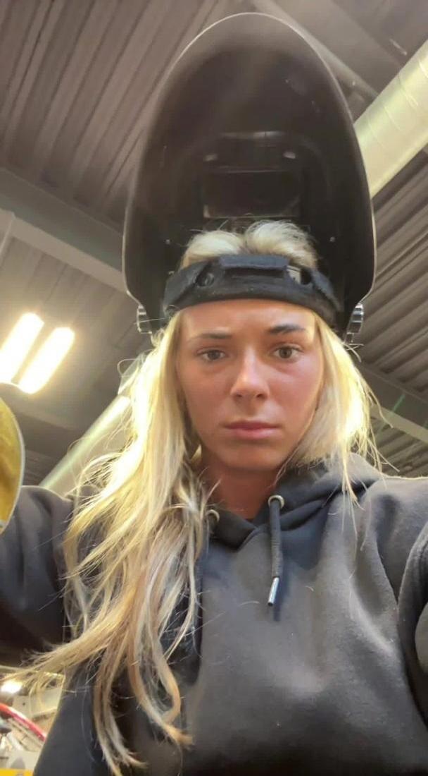A young woman with blonde hair, wearing a black hoodie and a welding helmet pushed up on her head, looks directly at the camera with a serious expression in what appears to be a workshop setting.