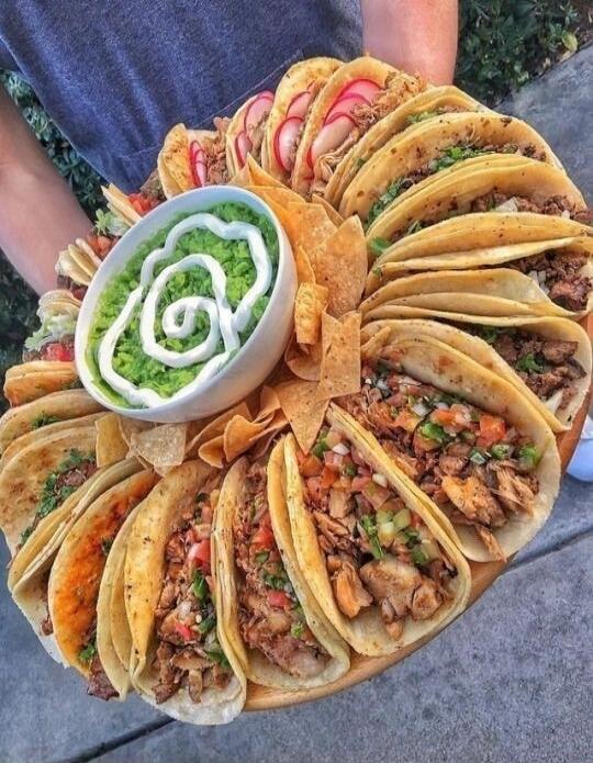 A large platter filled with various tacos, a bowl of guacamole, and tortilla chips.
