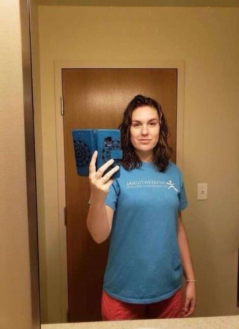 A woman takes a mirror selfie. She is wearing a blue t-shirt with the text 'INTERNATIONAL STUDENT ASSOCIATION' (reflected) and red shorts. She holds a phone in a blue case with a mandala pattern.