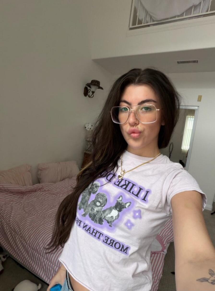 A young woman with long dark hair and glasses is taking a selfie, making a kissy face. Her t-shirt reads: 