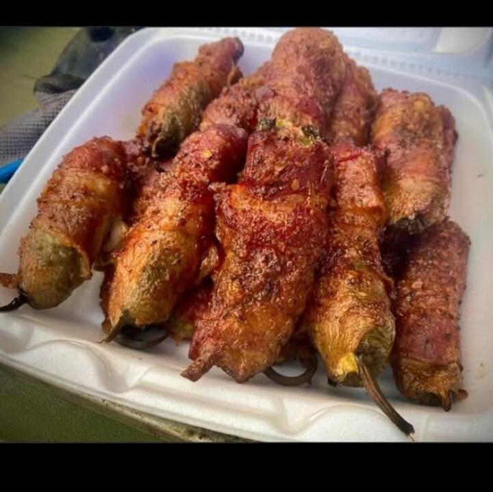 A close-up shot of several bacon-wrapped jalapeño poppers or stuffed peppers, golden brown and crispy, served in a white styrofoam container.
