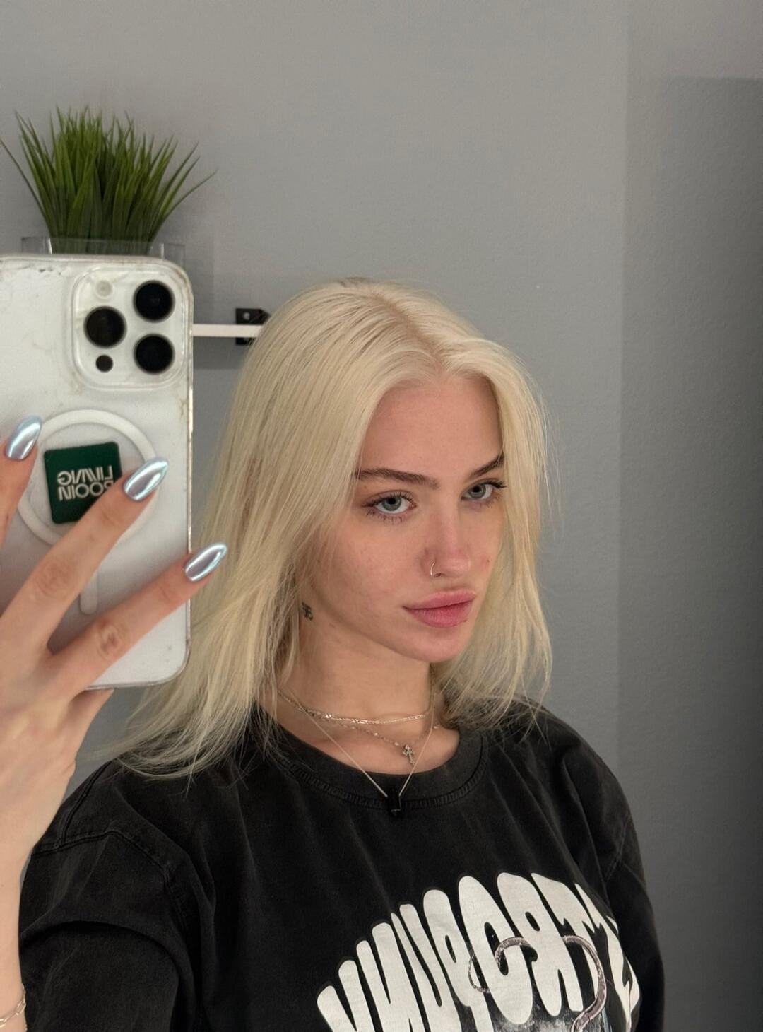 A young woman with long blonde hair and blue eyes takes a mirror selfie. She has a nose piercing and multiple necklaces. Her nails are painted with a metallic finish. She is wearing a dark t-shirt with some white graphic text. A small green plant is visible in the background.