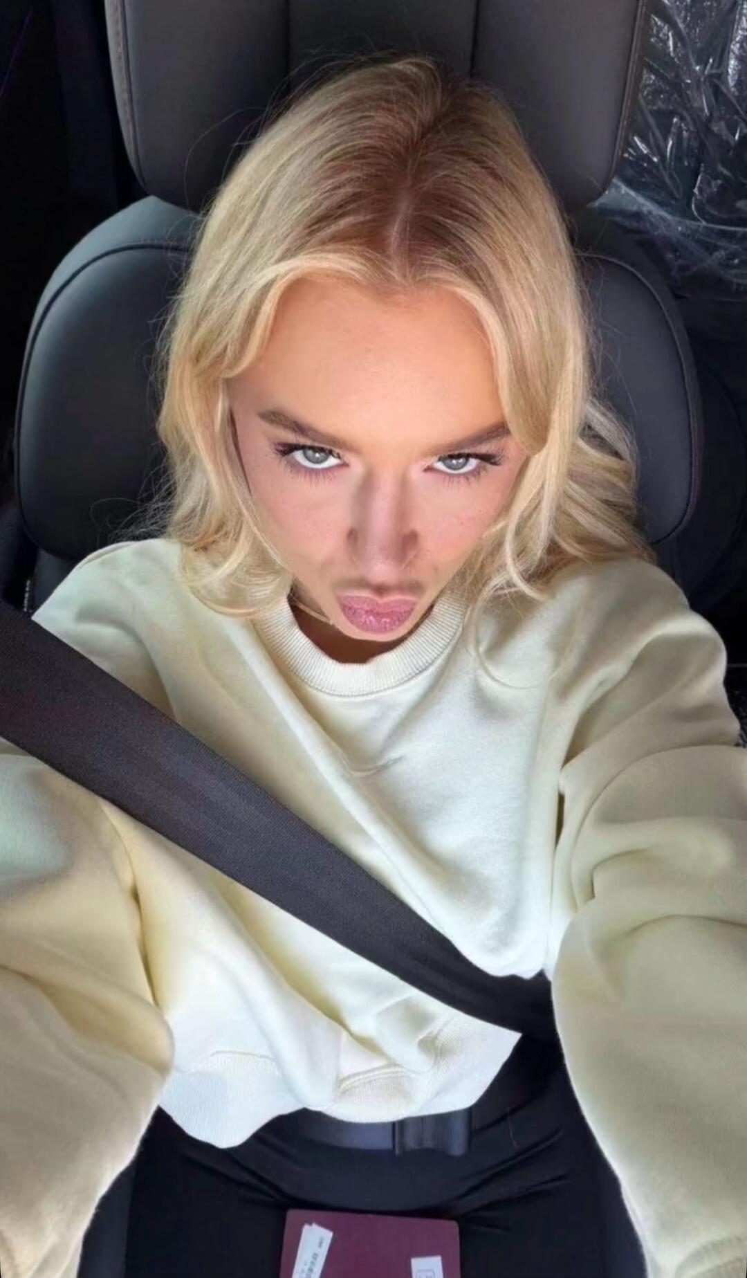 A blonde woman in a car, wearing a light sweatshirt and a seatbelt, looks at the camera with a playful, pouting expression and raised eyebrows. A document is visible on her lap.