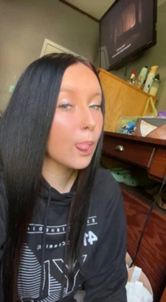 A young woman with long dark hair and light eyes sticks her tongue out playfully in a selfie.
