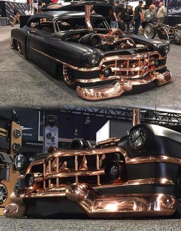 A highly customized classic car, possibly a Cadillac, with a matte black finish and extensive copper or rose gold accents on the grille, bumpers, engine components, and exhaust pipes. The vehicle is lowered and displayed at what appears to be a car show or exhibition, with people in the background.