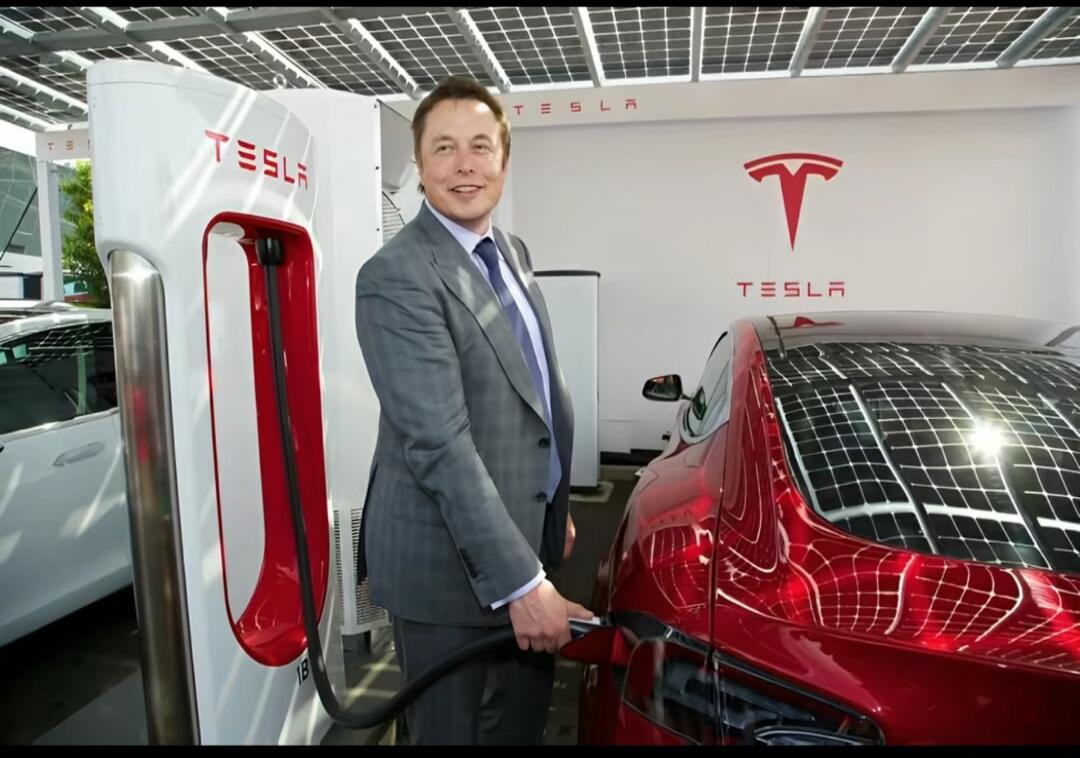 Elon Musk is seen smiling while plugging in a red Tesla car at a Tesla Supercharger station. The Tesla logo is visible on the charging station and the wall.