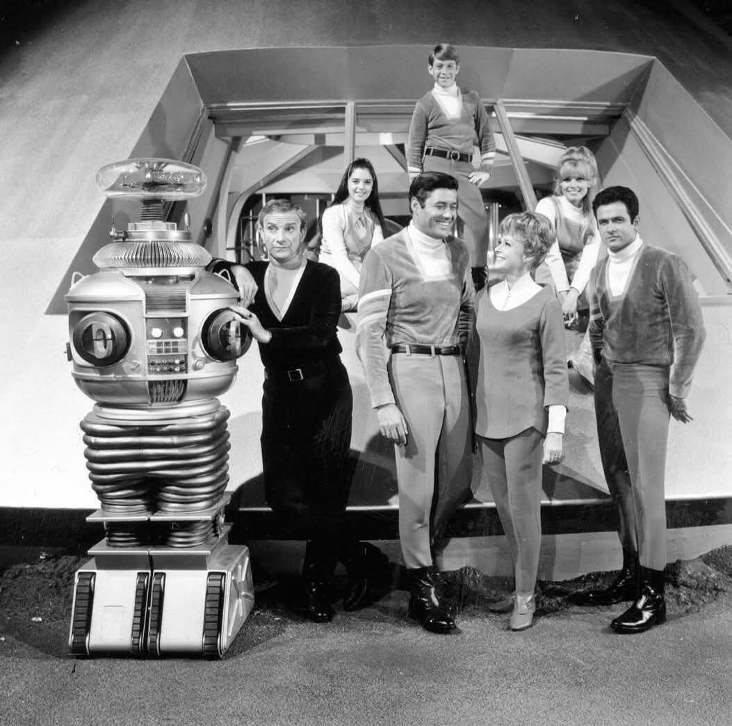 A black and white photo of the cast of the classic science fiction TV show Lost in Space, featuring seven actors and the iconic Robot B-9. They are posed in front of a futuristic structure, likely a spaceship.