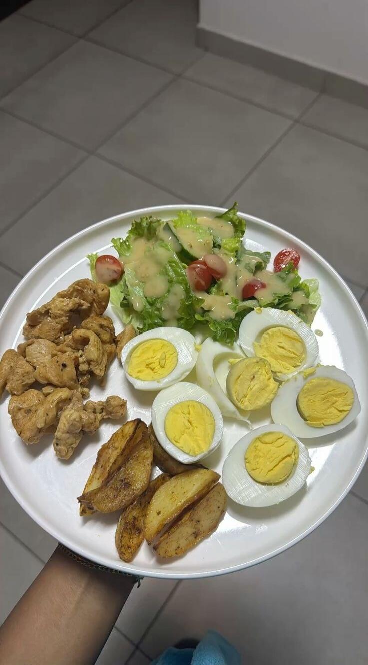 Plate with a mixed meal: salad with lettuce, cucumber, and tomatoes; hard-boiled eggs (sliced); fried chicken pieces; potato wedges.