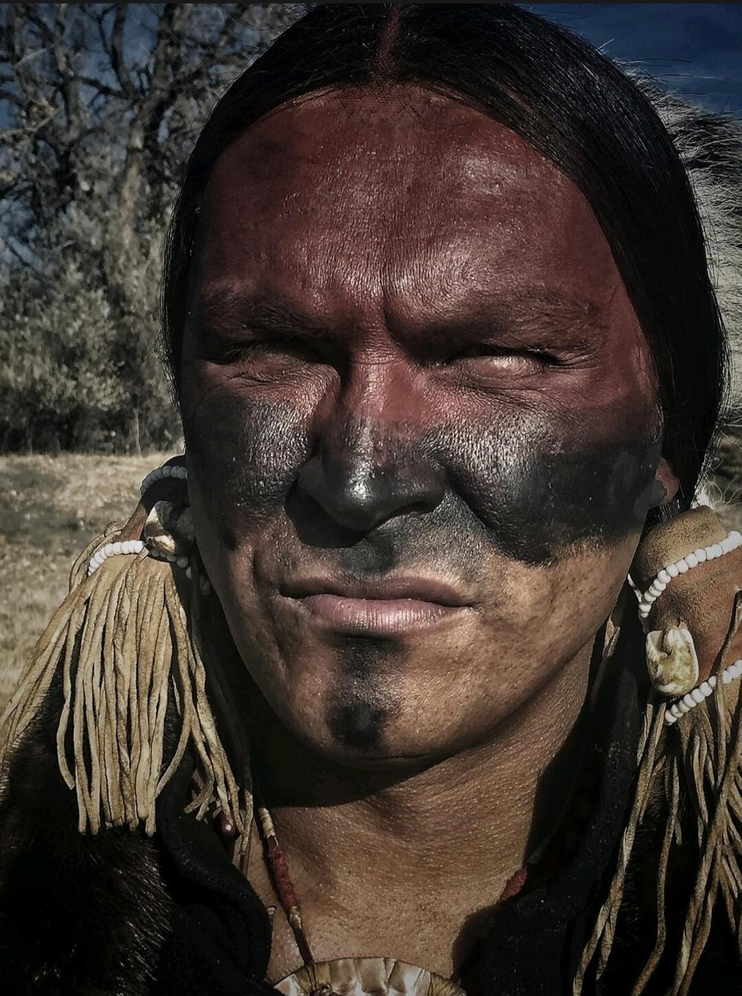 A portrait of a person with dark facial paint, long black hair, and traditional beaded adornments.