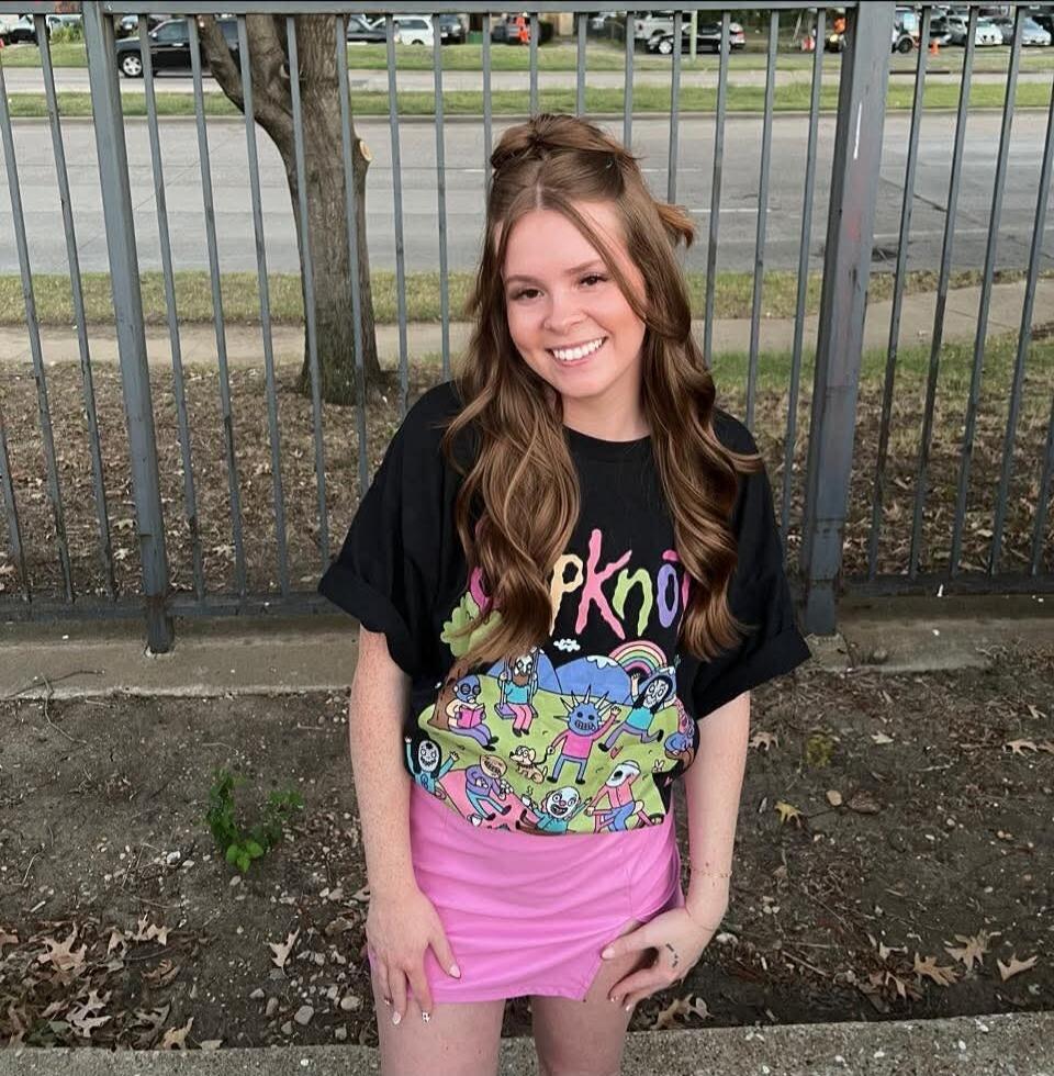 A young woman smiling, wearing a black t-shirt with 'pkno' and a colorful cartoon graphic, and a pink mini-skirt, standing outdoors in front of a metal fence.