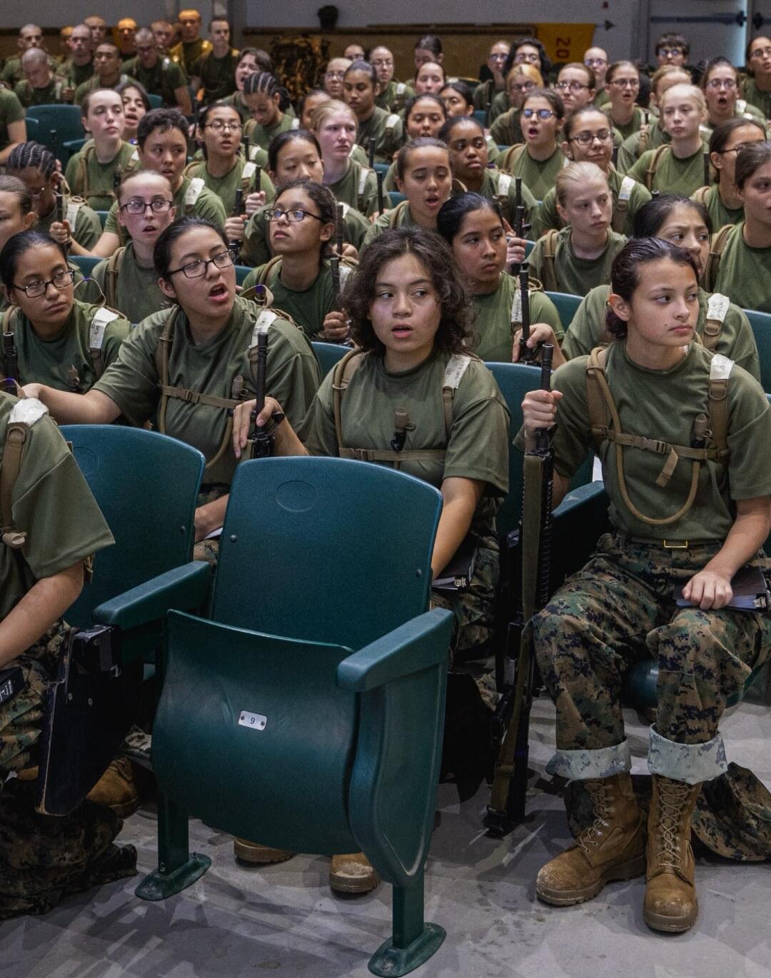 A large group of young women in military-style uniforms are seated in an auditorium, holding what appear to be training rifles. They are looking forward with attentive expressions, some with open mouths as if speaking or listening intently.