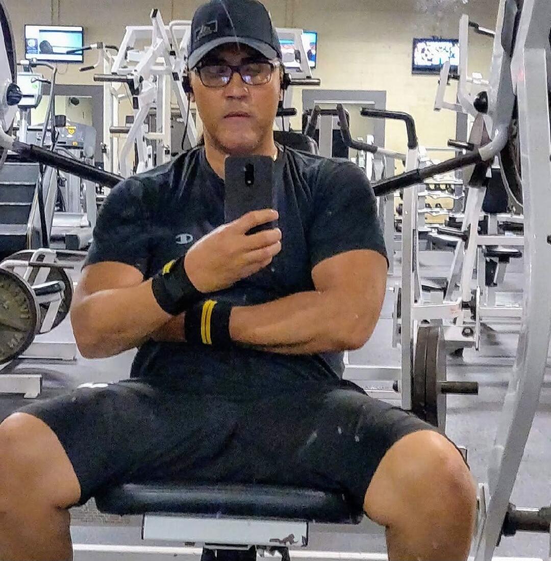 A man in a black t-shirt, black shorts, cap, and glasses is taking a mirror selfie in a gym. He is sitting on a bench, holding a smartphone, and wearing black and yellow wrist wraps. Various gym equipment is visible in the background.