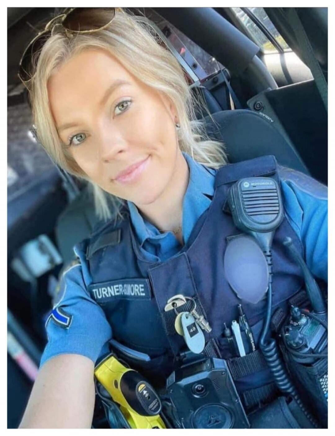 A blonde female police officer in uniform and a bulletproof vest takes a selfie inside a police car. Her vest has 'TURNER&MORE' written on it. A Motorola radio, a taser, and a body camera are visible. The car's interior shows 'AIRBAG'.