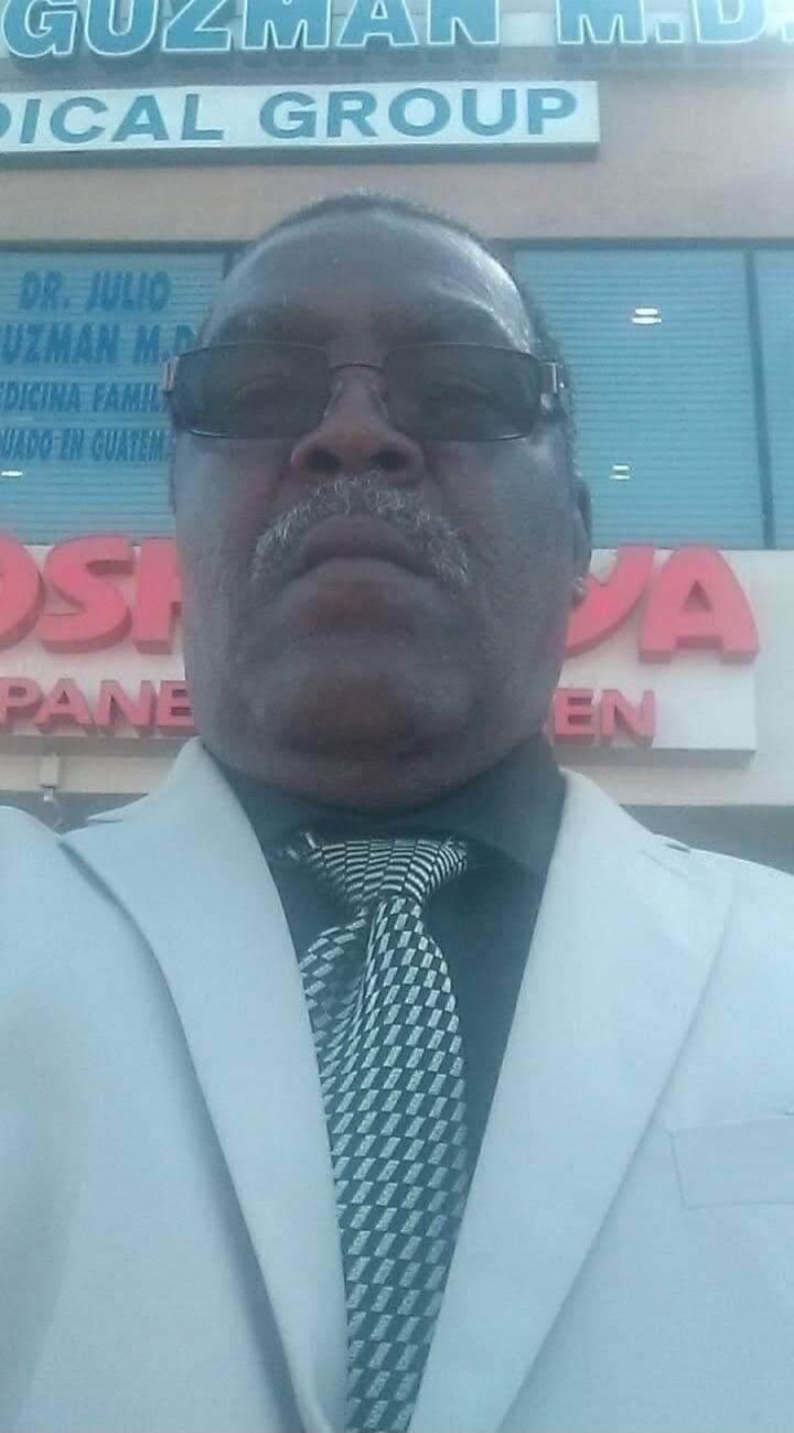 A man in a white suit jacket, dark shirt, and patterned tie, wearing sunglasses and a mustache, takes a selfie. In the background, signs for a medical office are visible, including 'GUZMAN M.D.', 'MEDICAL GROUP' (English), and 'DR. JULIO GUZMAN M.D.', 'MEDICINA FAMILIAR' (Spanish for Family Medicine). The word 'EN' is also visible.