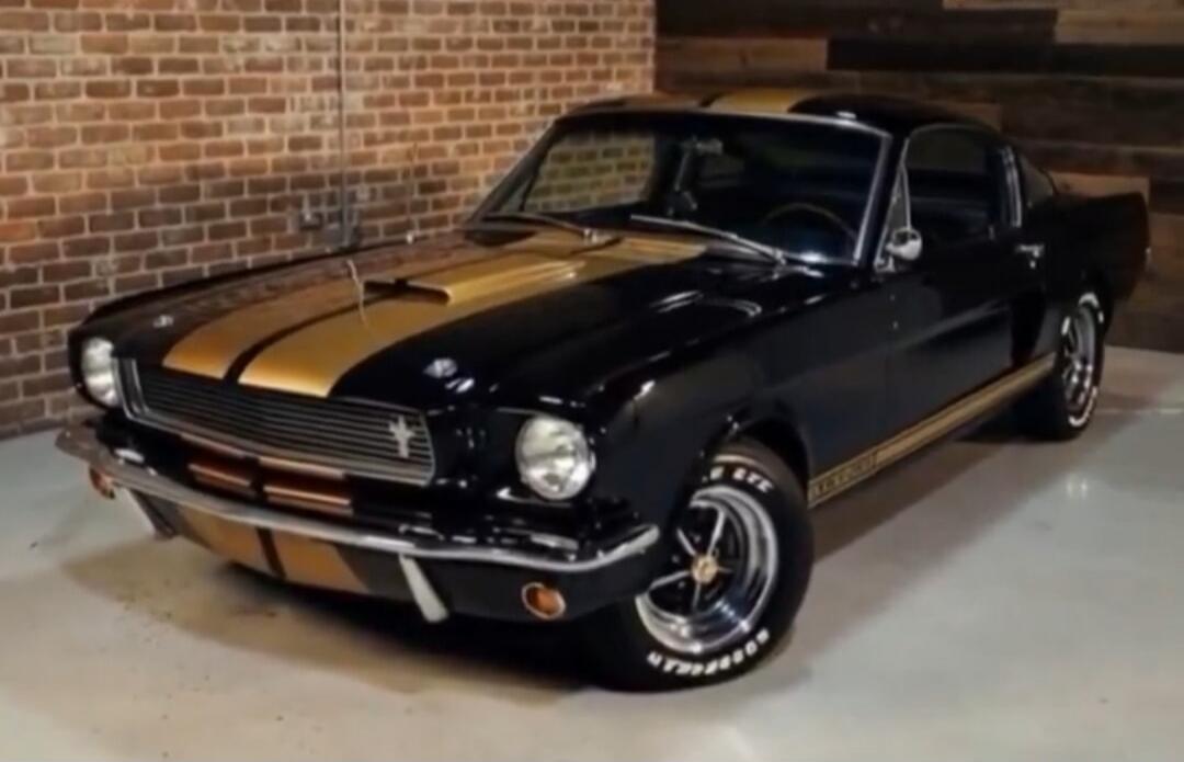 Black vintage car with gold racing stripes parked indoors next to a brick wall.