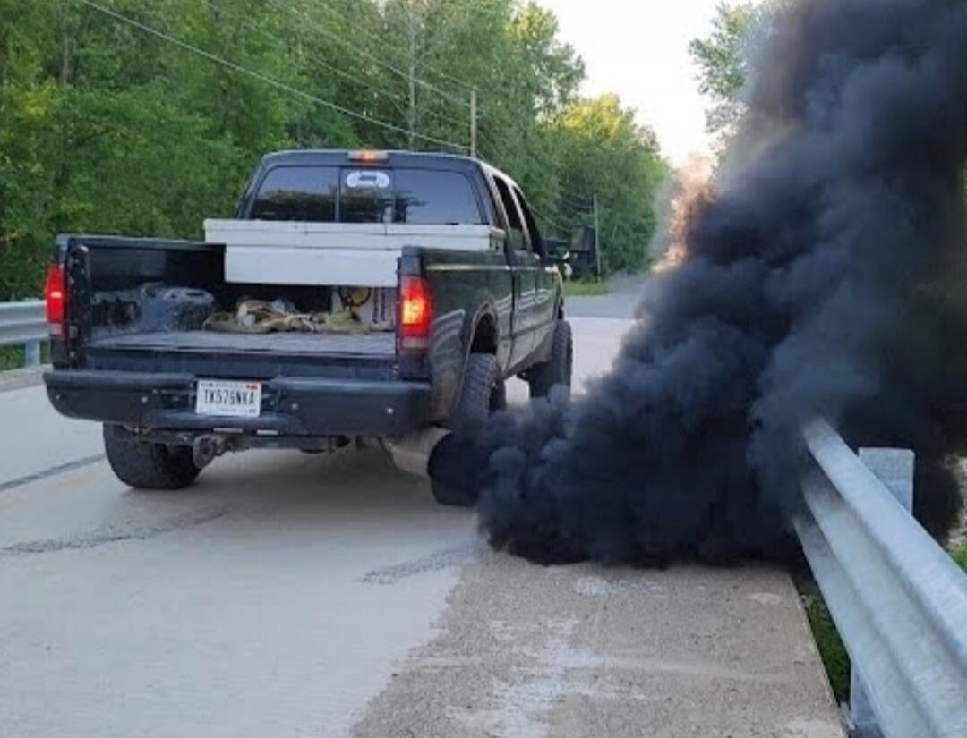 A dark-colored pickup truck on a road with thick black smoke coming from near the guardrail; the tailgate is open.