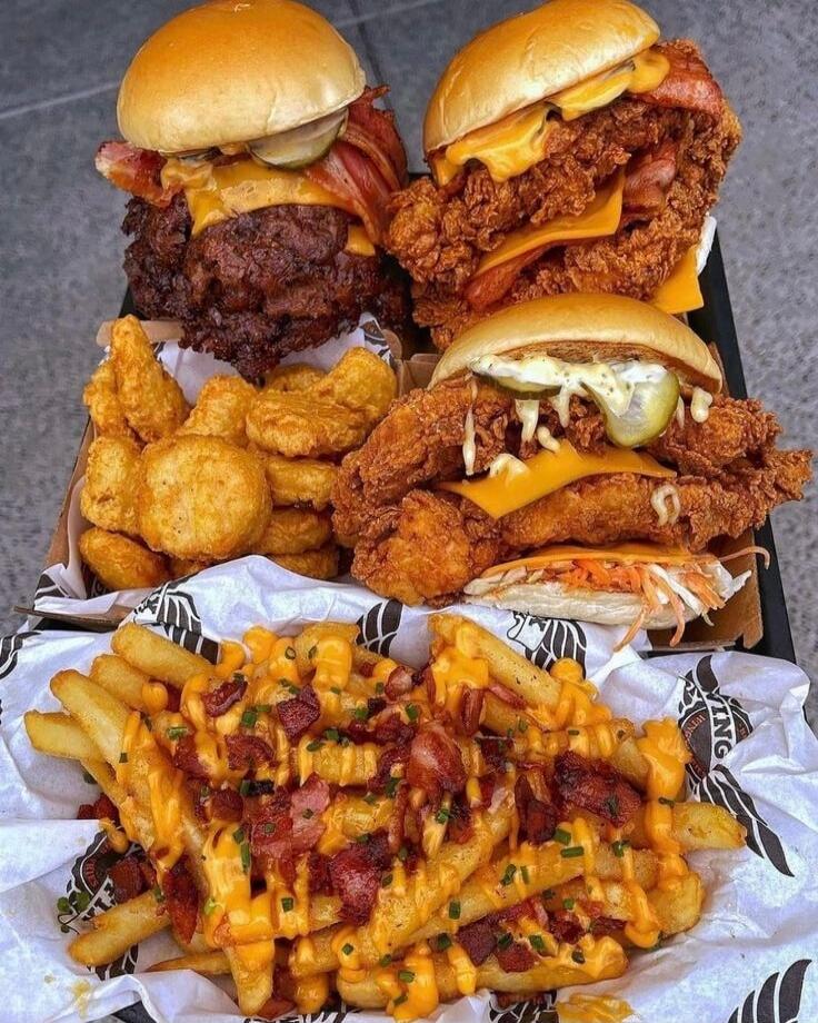 A tray of indulgent fast-food: two or three large fried chicken sandwiches with cheddar cheese and toppings like bacon and pickles, plus a pile of fried onion rings and chicken nuggets. In the foreground, loaded cheese fries topped with bacon bits and chives. Overall, a heavy, calorie-dense feast of fried comfort foods.