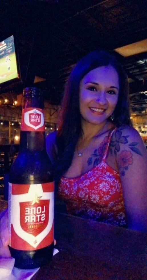 A smiling woman is holding a bottle of Lone Star beer. The label on the bottle clearly shows 'LONE STAR'. In the background, a television screen displays what appears to be a sports game.
