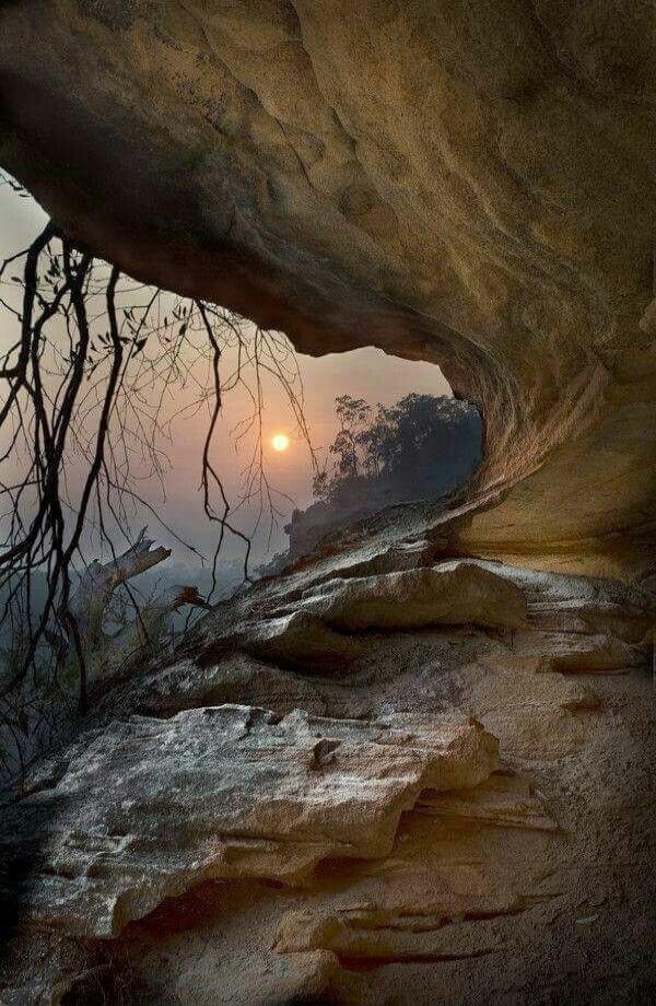 Sunset seen through a cave opening with rocky ledges and bare branches silhouetted against the hazy sky.