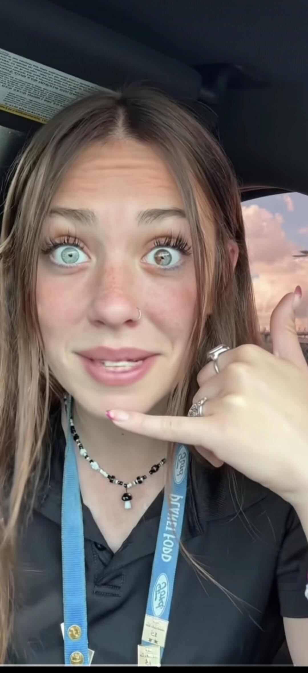 A young woman with heterochromia (one blue eye, one brown eye) making a 'call me' gesture. She is wearing a black polo shirt and a blue lanyard with 'Ford' and 'PLANET FOOD' text. She also has a mushroom necklace.