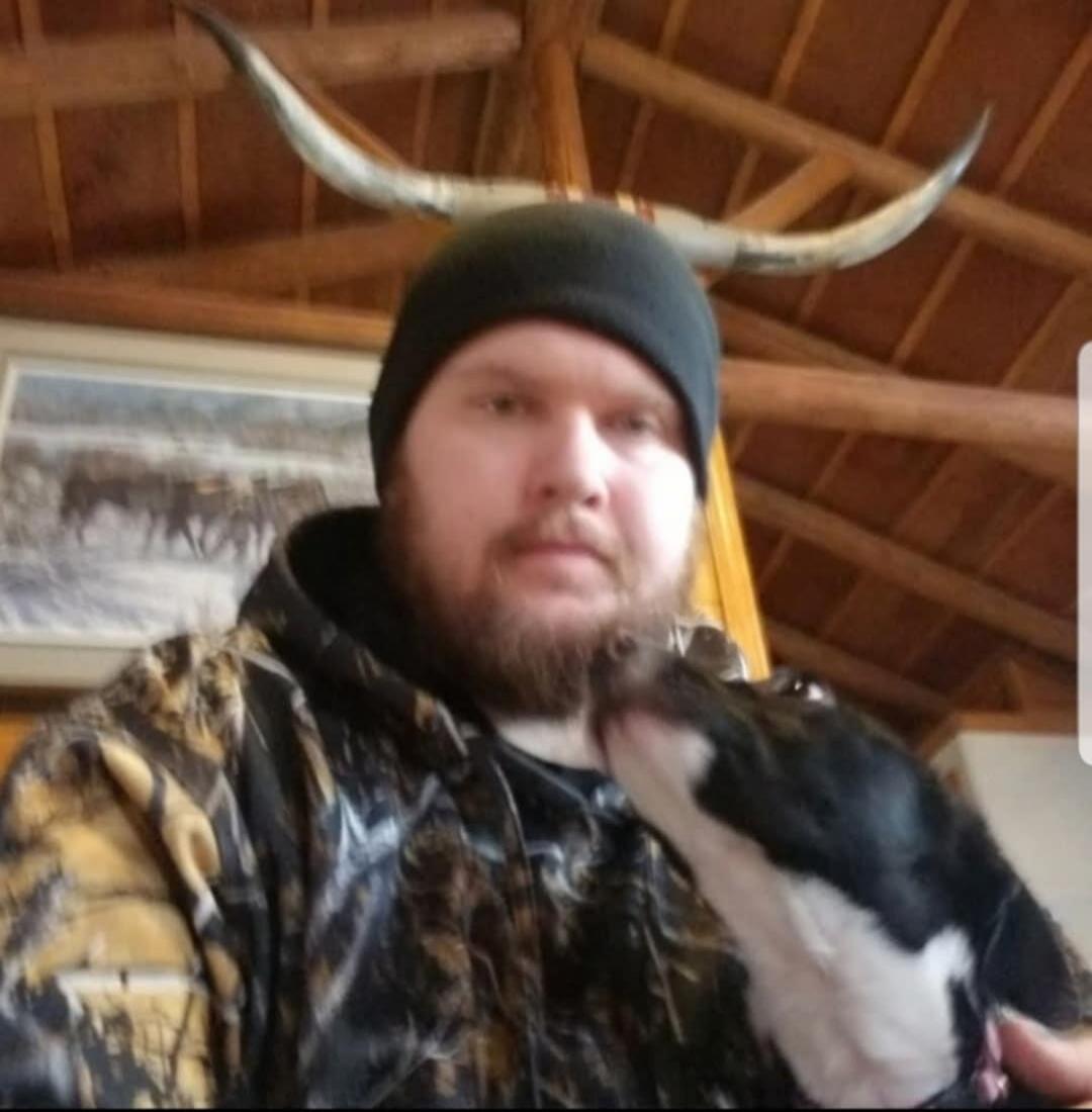 A man with a beard and a beanie is holding a small black and white dog that is licking his face. In the background, there are wooden beams and longhorn horns.