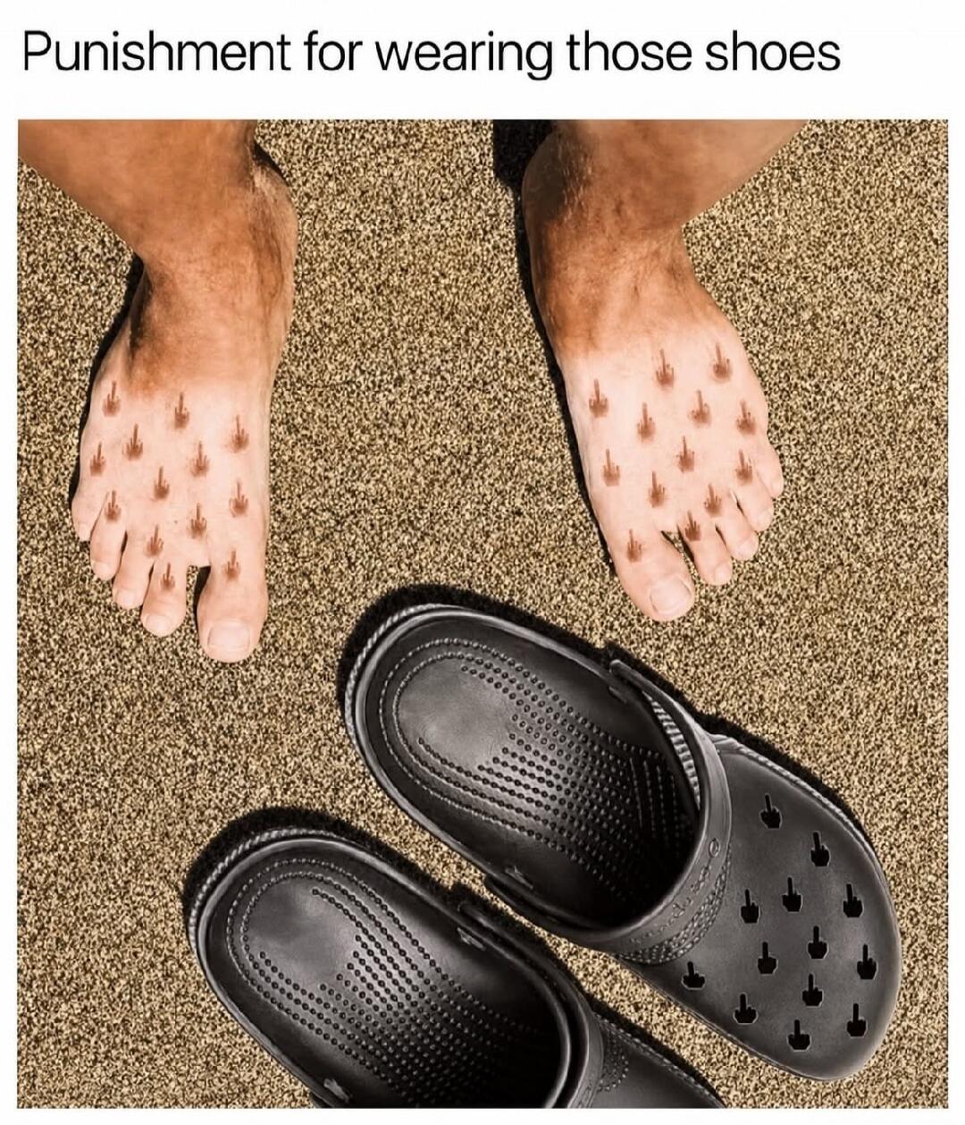 Punishment for wearing those shoes. A person's feet show tan lines in the shape of multiple middle fingers, matching the pattern of holes on a pair of black Crocs shoes lying on the sand.