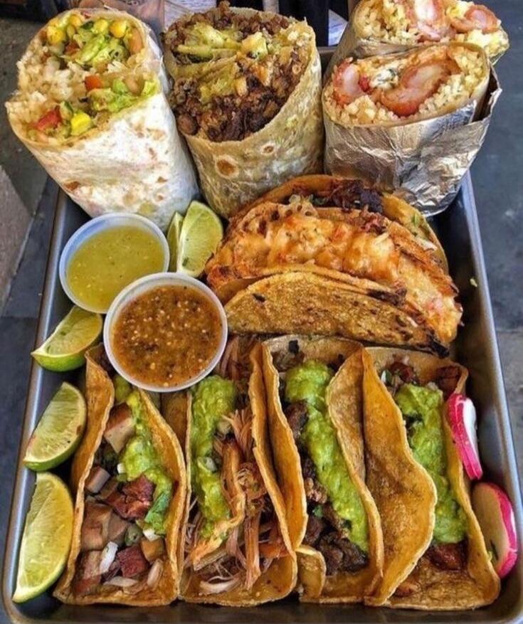 Assorted tacos and burritos with sauces and lime wedges.