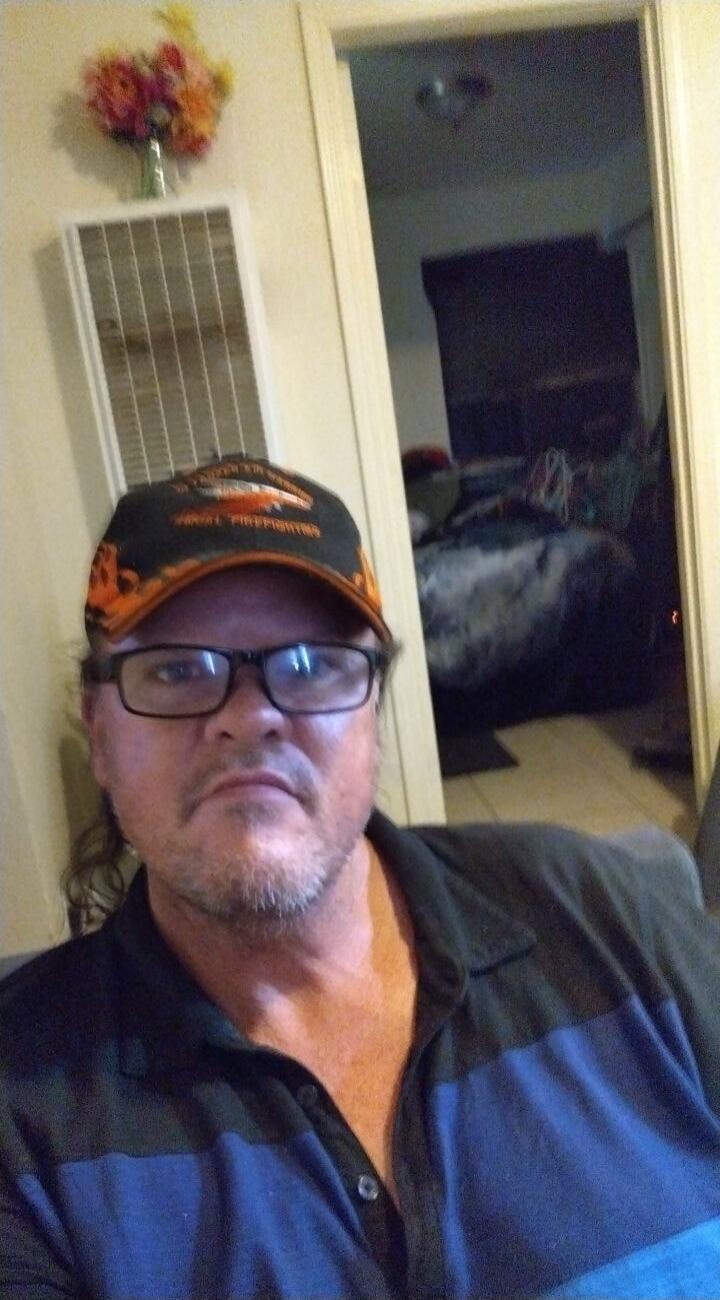 A man wearing a black and orange cap with 'FINAL FIREFIGHTERS' text and glasses, taking a selfie indoors.