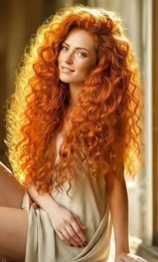 A woman with long curly red hair posing indoors.