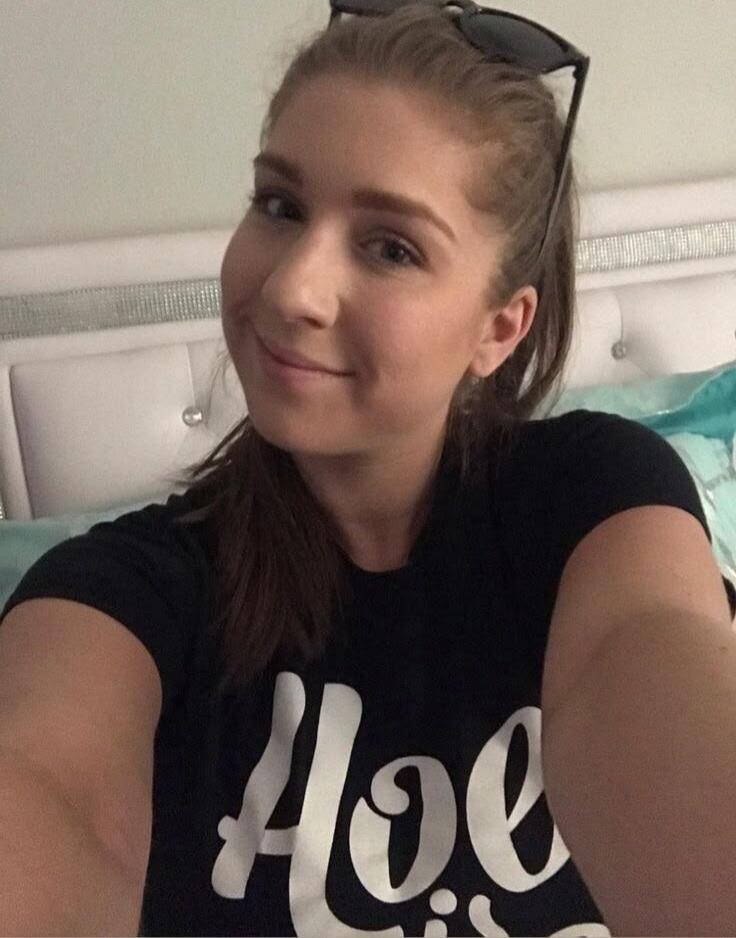 A young woman takes a selfie, smiling. She is wearing a black t-shirt with the word 'Hoe' printed in white. Sunglasses are on her head.