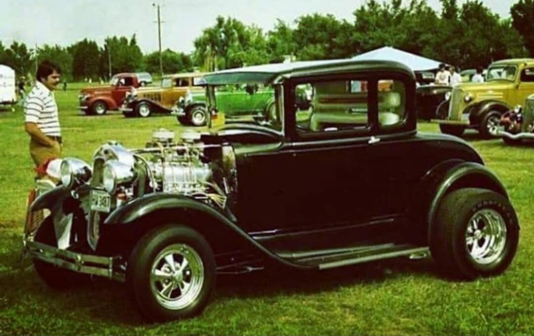 A classic black hot rod car with a prominent engine, possibly a supercharger, is parked on a green grassy field. A man in a striped shirt and khaki pants stands next to it, observing the vehicle. In the background, other vintage cars are visible under a clear sky with trees. The license plate reads 'PW 347'.