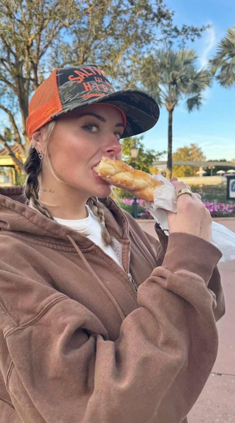 Woman in a brown hoodie and an orange/black cap is biting into a pastry outside.