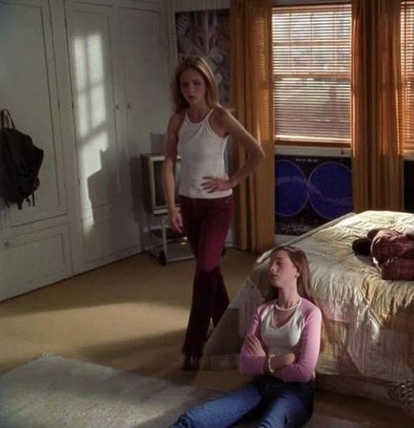 A woman stands in a bedroom with her hand on her hip, looking down at another woman sitting on the floor with her eyes closed and arms crossed, leaning against a bed.