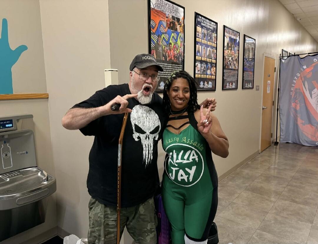 An older man with a white beard and glasses, wearing a black t-shirt with a Punisher skull logo and a cap that says 'FOGMA', poses with a woman. The woman has dark skin and dreadlocks, wearing a green and black wrestling-style outfit that says 'Sassy Assassin KILLA JAY'. She is making a peace sign. The man has an exaggerated expression and is holdi