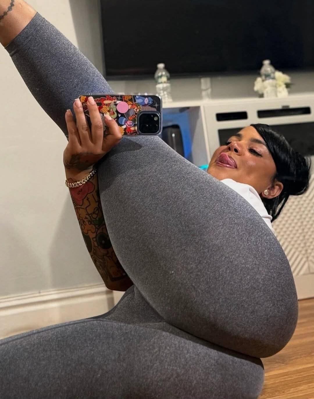 A woman wearing gray leggings sits on the floor, taking a selfie with her phone while stretching her leg up.
