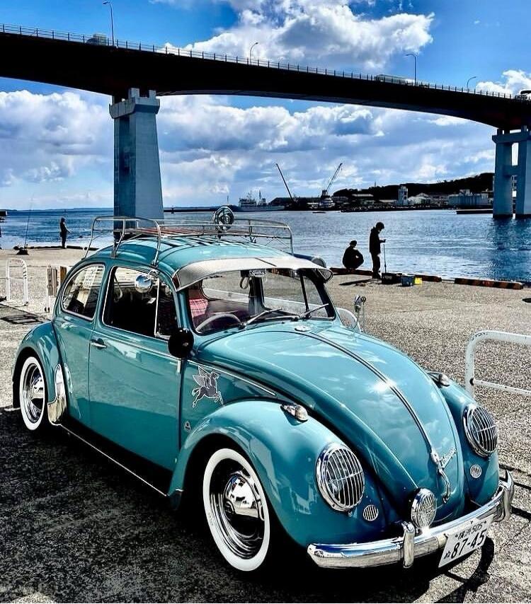 A turquoise vintage Volkswagen Beetle with whitewall tires parked on a beach, with a blue bridge in the background. The car has chrome accents and a roof rack. License plate visible on the front.